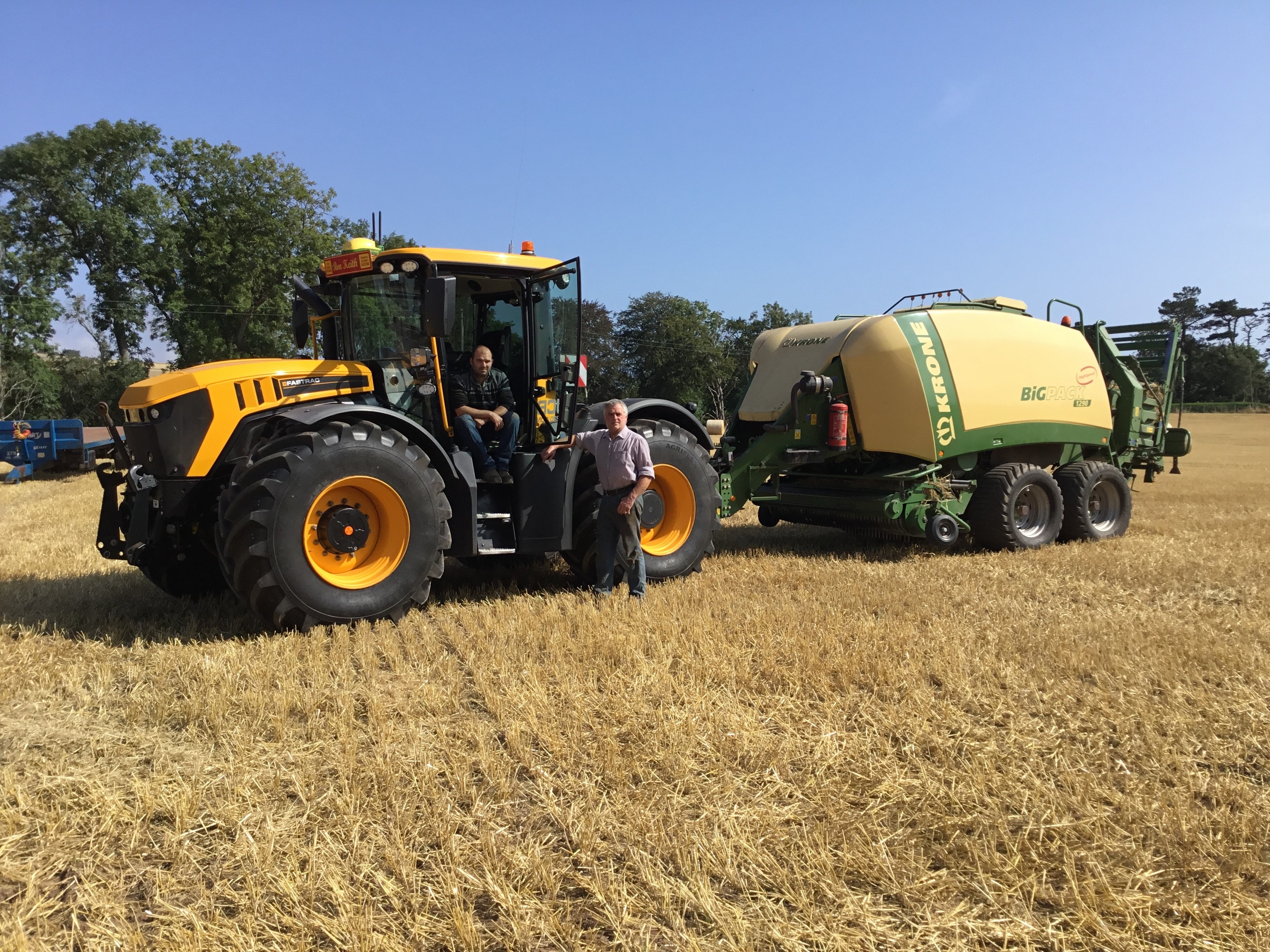 Two people standing beside a JCB Fastrac tractor and baler in a harvested field.